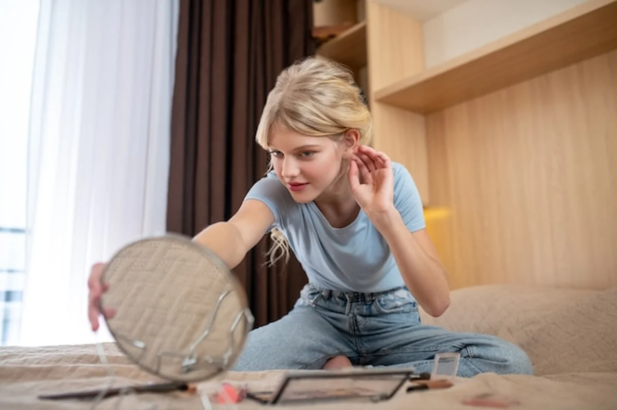 Dziewczyna w niebieskiej koszuli siedząca na łóżku patrząca w lustro