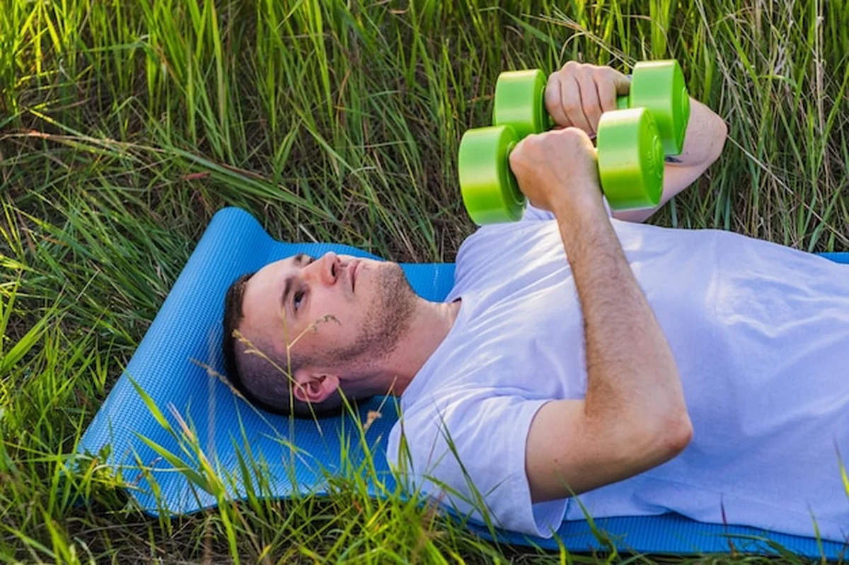 Mężczyzna ćwiczy z zielonymi hantlami na zewnątrz na niebieskiej matce na bujnym zielonym polu w ciągu dnia