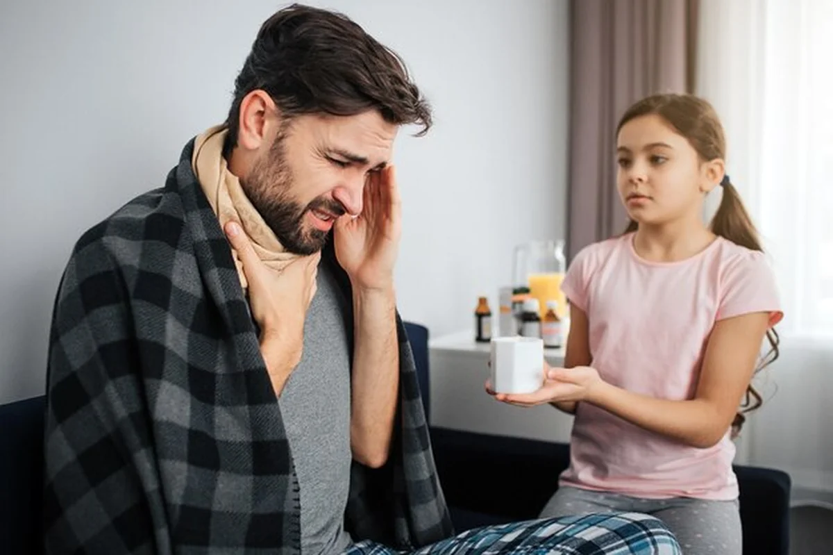 Młody chory mężczyzna siedzi z córką, trzymając ręce na gardle, a dziewczynka patrzy. Kubek z lekiem w ręce